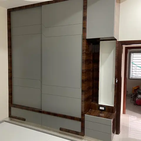 Bedroom sliding wardrobe in matte grey with a dark wood-veneer frame, paired with a built-in dresser unit and open shelf niche beside the doorway.