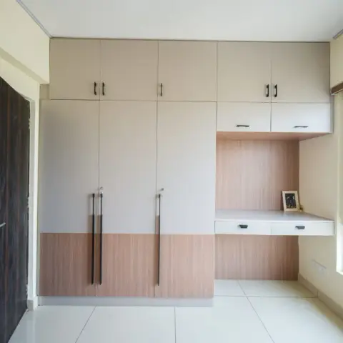 Bedroom storage wall in beige and oak laminate with black bar handles, the wardrobe run turning the corner into a wall-hung two-drawer study ledge under the