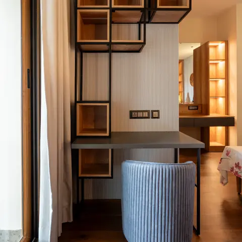 Bedroom study nook with a black metal-frame open shelving unit over a fluted-panel desk backsplash and a ribbed blue barrel chair, with the bed visible beyond.