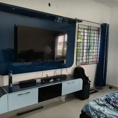 Bedroom TV wall in glossy navy and light-blue laminate with a floating panel, navy curtains, and the edge of a floral-print bed in the foreground.