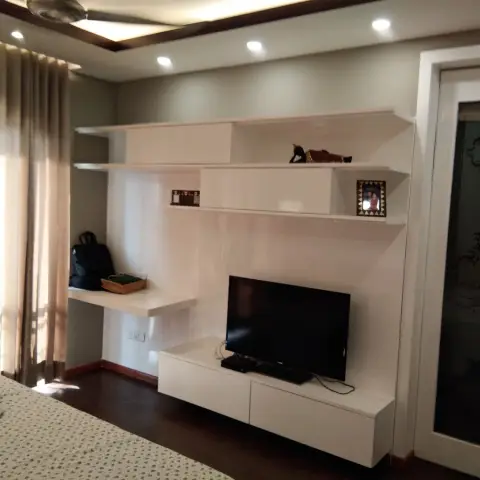 Bedroom TV wall in white lacquer - a wall-hung console with staggered open shelves and a small floating desk ledge to its left, beside an etched-glass door