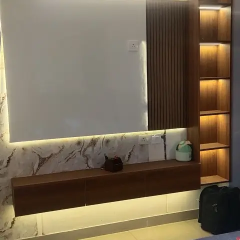 Bedroom TV wall with a backlit recess framed in beige marble-print cladding, a floating walnut console below and a lit open-shelf tower to one side