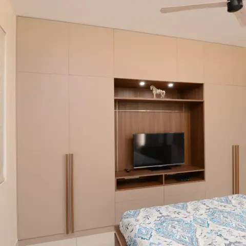Bedroom wall built out as a floor-to-ceiling beige wardrobe run with lofts, wrapping a walnut-veneer TV niche lit by two small spots with an open drawer