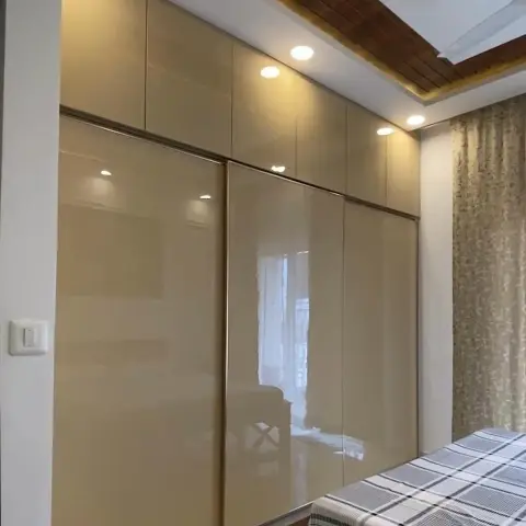 Bedroom wall taken up by a beige high-gloss sliding wardrobe with separate loft shutters, under a false ceiling with an inset timber-slat panel and cove light
