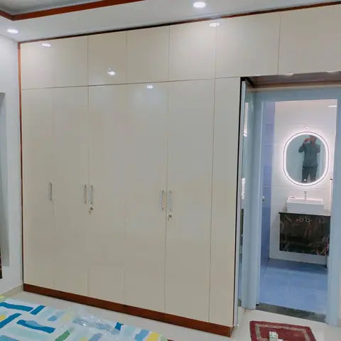 Bedroom wardrobe wall in glossy cream with wood-trim edging, opening onto an ensuite bathroom with a marble vanity and backlit oval mirror.