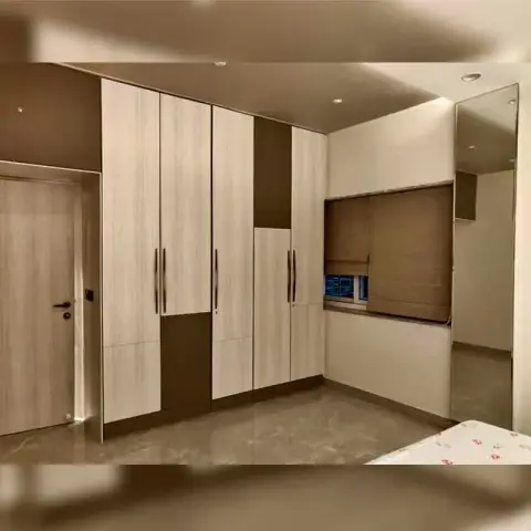 Bedroom wardrobe wall in pale ash laminate broken by dark brown inset bands, running up to a built-in window seat under a brown roman blind with a full-height