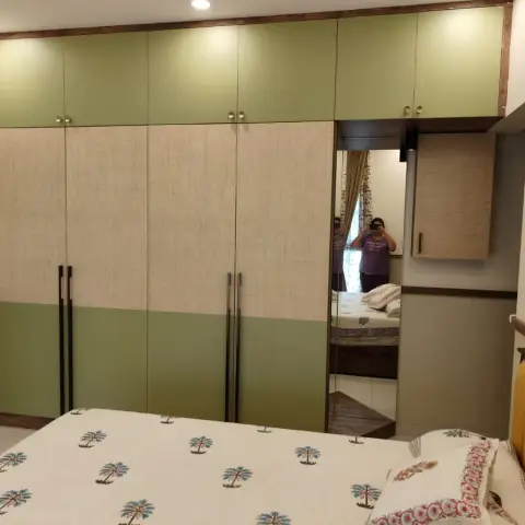 Bedroom wardrobe wall in sage-green and jute-textured laminate with separate lofts above and a full-height mirror panel at the end