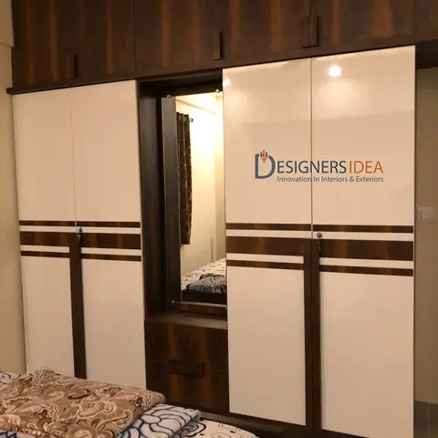 Bedroom wardrobe wall in white-and-walnut laminate with a built-in mirror and dressing drawers, printed bedcover in the foreground.