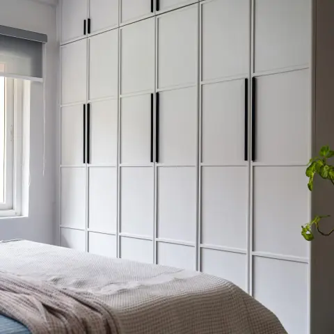 Bedroom wardrobe wall in white shaker-profile shutters running floor to ceiling with a separate loft tier and slim matt-black bar handles
