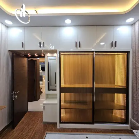 Bedroom wardrobe wall of amber reeded-glass sliding shutters lit from inside, white gloss loft cabinets