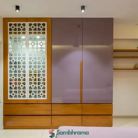 Bedroom wardrobe wall with a wood-framed CNC jaali insert door, glossy mauve lacquer shutters, open display shelves, and warm cove ceiling lighting.