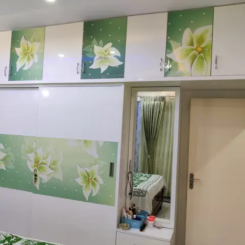 Bedroom wardrobe wall with floral-print glossy sliding doors in green and white, a built-in dressing mirror unit, and a sliver of bed visible in the foreground.