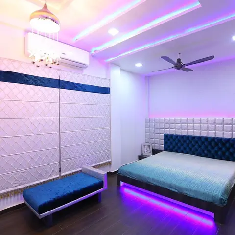Bedroom washed in pink-and-blue RGB cove light, with a navy tufted-velvet headboard over silver quilted upholstered panels and an LED strip glowing out from