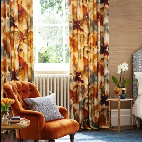 Bedroom window corner dressed in a large-scale watercolour-floral drape, with a rust velvet buttoned armchair on a faded blue rug and a white orchid on a gilt