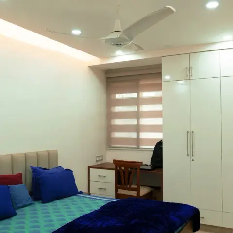 Bedroom with a beige channel-tufted headboard and royal-blue bedding, a white four-shutter wardrobe run with loft units filling the far wall and a small wooden