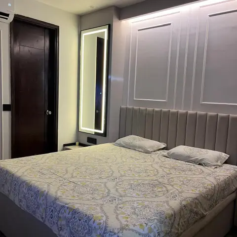 Bedroom with a channel-tufted greige headboard against a grey wall of white moulding panels lit from a concealed strip