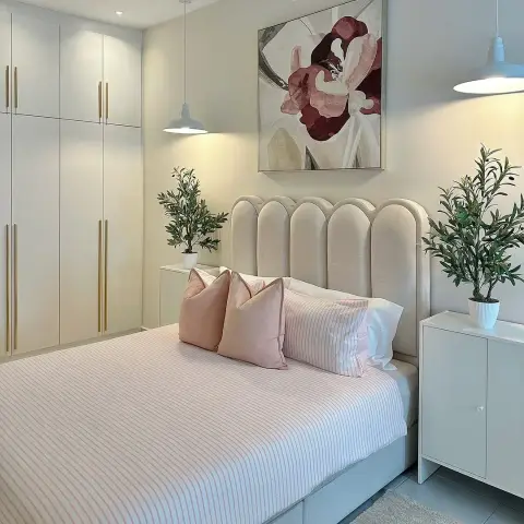 Bedroom with a cream scalloped-arch upholstered headboard and blush striped bedding, flanked by two white dome pendants and potted olive trees
