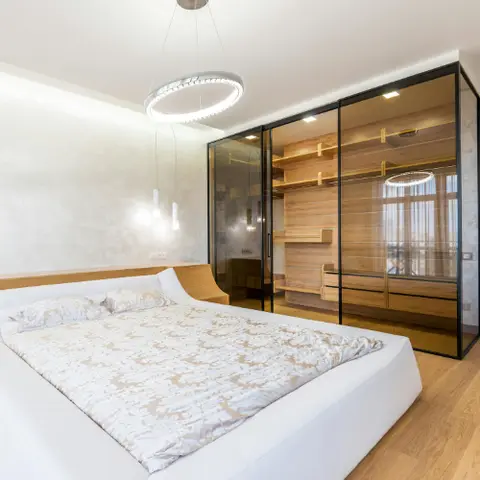 Bedroom with a curved white platform bed beneath a double-ring LED pendant chandelier, beside a black-framed glass walk-in wardrobe with wood-slat shelving.