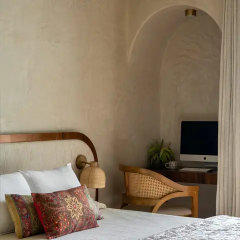 Bedroom with a curved wood-and-linen headboard and block-print cushions, opening to an arched lime-plaster niche with a built-in study desk and cane chair.