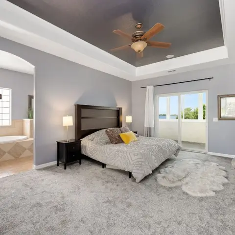Bedroom with a dark tray ceiling and grey carpeting, opening through an archway to an en-suite bathroom with a soaking tub and chandelier.