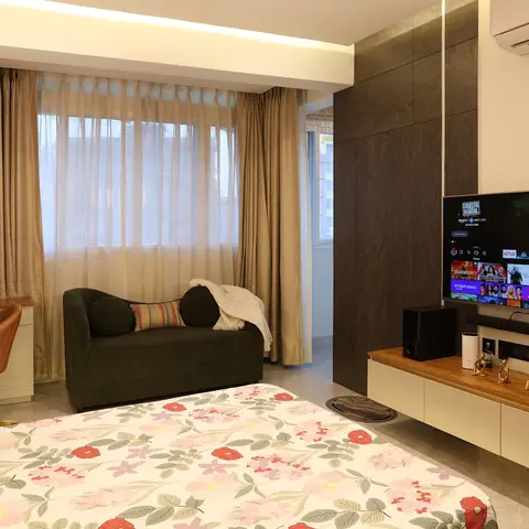 Bedroom with a floating wood-and-white TV console against a dark textured wardrobe wall, a study desk nook, and a velvet chaise lounge by the window.
