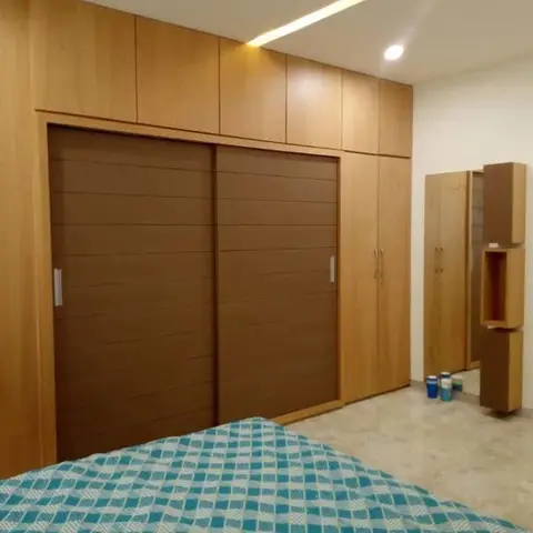 Bedroom with a floor-to-ceiling wood-laminate sliding-door wardrobe under a lit ceiling cove, seen from the foot of a teal-checked bed.