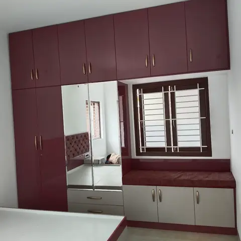 Bedroom with a floor-to-ceiling wine-maroon glossy wardrobe with a mirrored shutter, and a cushioned window seat over white cabinetry.