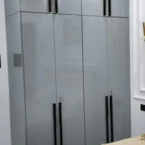 Bedroom with a floor-to-ceiling glossy grey wardrobe with black vertical pull handles and cove ceiling lighting, bed in foreground.