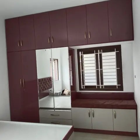 Bedroom with a floor-to-ceiling maroon high-gloss wardrobe with a mirrored shutter, and a built-in cushioned window seat with storage cabinets below.