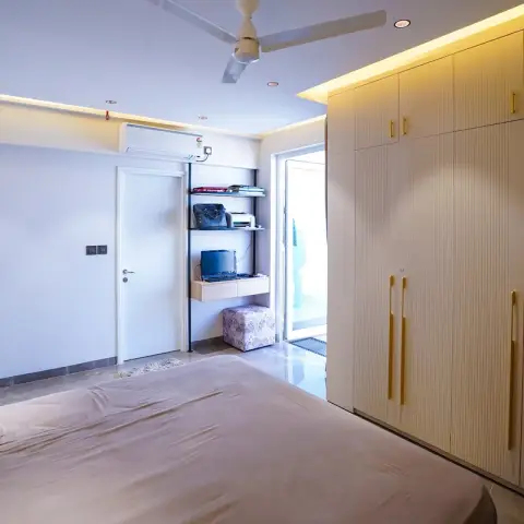 Bedroom with a floor-to-ceiling fluted cream wardrobe with brass pull handles and a cove-lit ceiling, small floating study shelf beside the balcony door