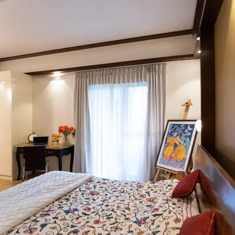 Bedroom with a floral embroidered bedspread on a dark wood bed, a glossy white wardrobe, a small writing desk by the window