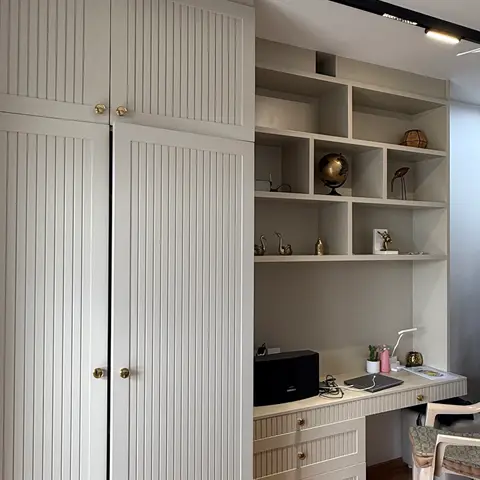 Bedroom with a fluted-panel wardrobe and an open shelving-and-study-desk unit styled with curios, brass hardware throughout, bed visible in the foreground.