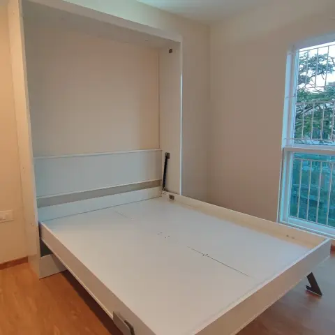 Bedroom with a folding wall-mounted Murphy bed pulled down, unmade and unstyled, wood-look flooring and a barred window.