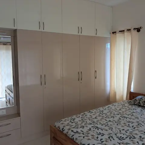Bedroom with a full-height glossy beige wardrobe wall spanning the room, a built-in dresser with mirror, and a wood bed dressed in a floral-print bedspread.