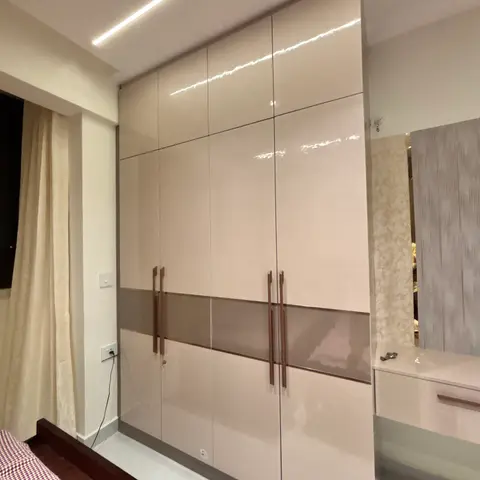 Bedroom with a full-height glossy beige wardrobe featuring a contrasting taupe glass band and wood baton pull handles, beside a mirrored dressing table.