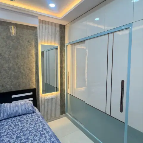 Bedroom with a full-height glossy white sliding wardrobe with teal-grey accent panels, a backlit mirror niche framed in wood-slat panelling