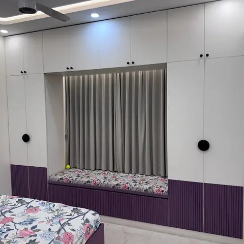 Bedroom with a full-height white wardrobe flanking a curtained window seat, fluted purple panelling at the base, and floral bedding matching the seat cushion.