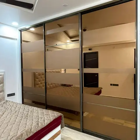 Bedroom with a full-wall bronze-tinted mirrored sliding wardrobe reflecting a tufted headboard bed on a wood-look tile floor.