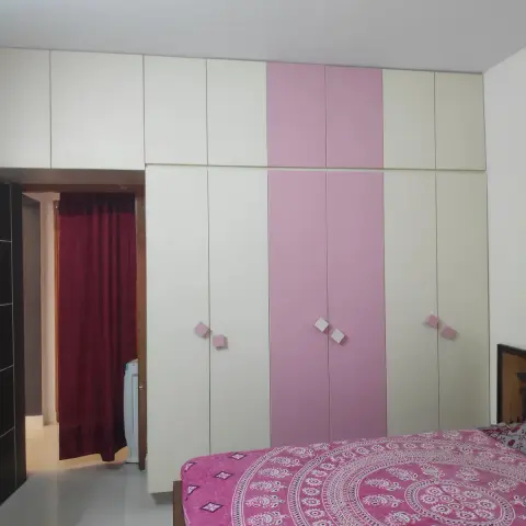 Bedroom with a full-wall cream-and-pink color-blocked wardrobe and a mandala-print pink bedspread, doorway to a maroon-curtained utility area beyond.