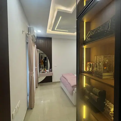 Bedroom with a glass-fronted, backlit wardrobe display for jewellery and bags, a round mirror set into dark wood fluted panelling, and a cove-lit false ceiling.