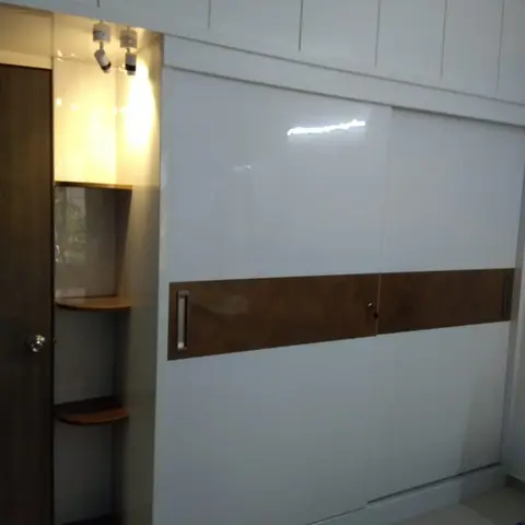 Bedroom with a glossy white sliding-door wardrobe featuring a wood-laminate accent band and a corner open shelf unit