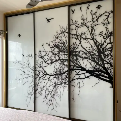Bedroom with a gold-framed sliding wardrobe whose glass shutters are printed with a black tree-and-bird silhouette, pink bedspread in the foreground.