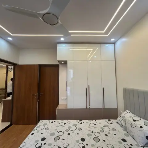 Bedroom with a grey fluted upholstered headboard beside a floor-to-loft high-gloss ivory wardrobe, under a false ceiling cut with continuous LED profile lines