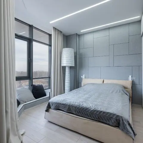 Bedroom with a grey upholstered geometric panelled headboard wall, linear cove ceiling lighting, a built-in window seat nook, and a sliding wardrobe.