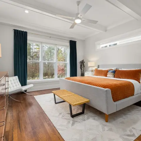 Bedroom with a grey upholstered king bed dressed in burnt-orange bedding, teal floor-length curtains and a mid-century-style tufted white lounge chair in the