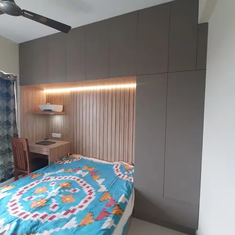 Bedroom with a grey wardrobe wall, a wood-slat headboard panel with concealed LED lighting, and a built-in study desk with a printer shelf.