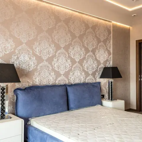 Bedroom with a large silver damask-pattern wallpaper accent wall, cove ceiling lighting, a blue velvet upholstered bed