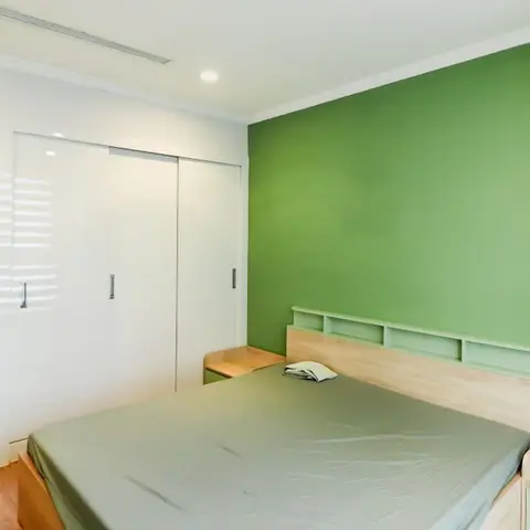 Bedroom with a leaf-green accent wall behind a maple-laminate bed whose headboard carries built-in open niches, facing a white gloss sliding wardrobe.