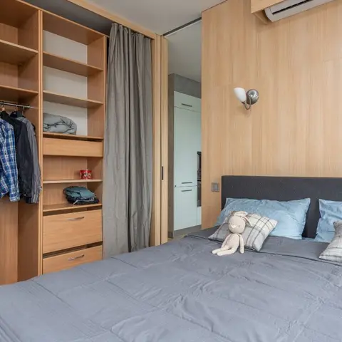 Bedroom with a light-oak veneer headboard wall, twin chrome globe sconces and an open shelved wardrobe half-hidden by grey drop curtains beside the bed.