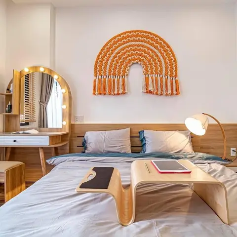 Bedroom with a macrame rainbow wall hanging above a wood headboard and an arched bulb-lit dressing mirror.